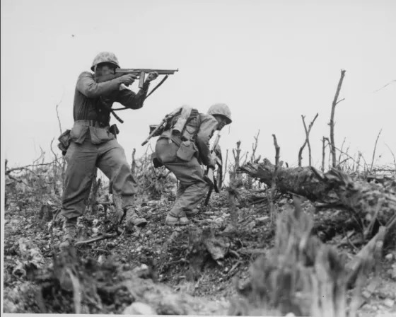 A Marine of the 1st Marine Division draws a bead on a Japanese sniper with his tommy-gun as his companion ducks for cover. The division is working to take Wana Ridge before the town of Shuri. Okinawa, 1945.